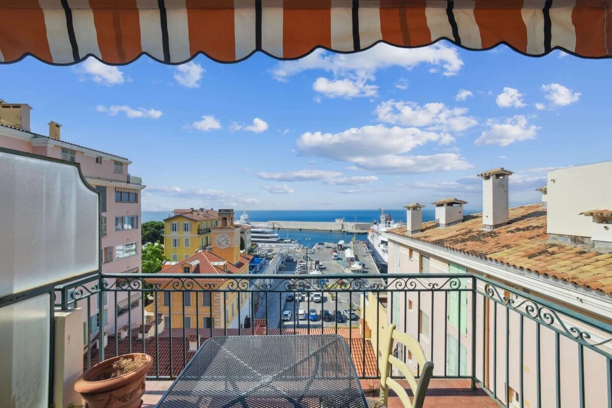 Balcony view of a vibrant coastal town with colorful buildings, a bustling port, and the blue Mediterranean Sea under a partly cloudy sky, framed by an orange awning.