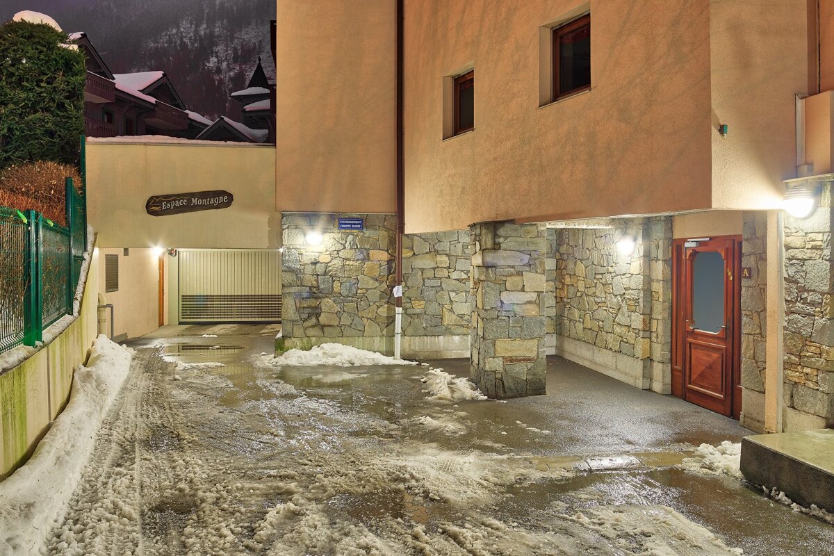 A snowy, slushy driveway at dusk, illuminated by lights, leading to a garage under buildings with stone bases and an 