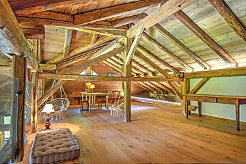 A spacious, rustic wooden loft with exposed beams, light wood floors, and cozy seating areas including hanging chairs and an ottoman.