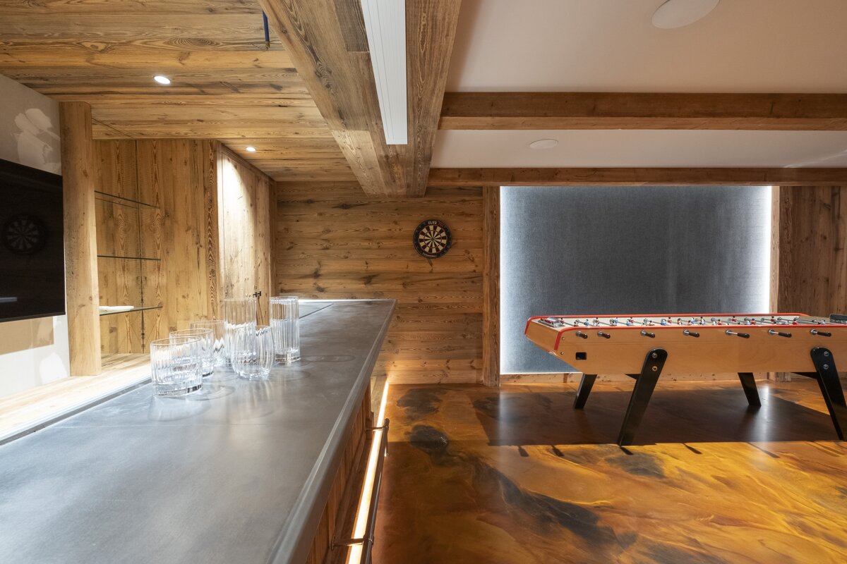 A wood-paneled game room with a sleek bar, foosball table, and dartboard. Features a striking gold-swirled floor and modern lighting.