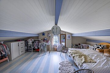 A light-filled attic bedroom with a blue and white striped floor, sloped paneled ceiling, a bed, vanity, and clothes racks, creating a cozy vibe.