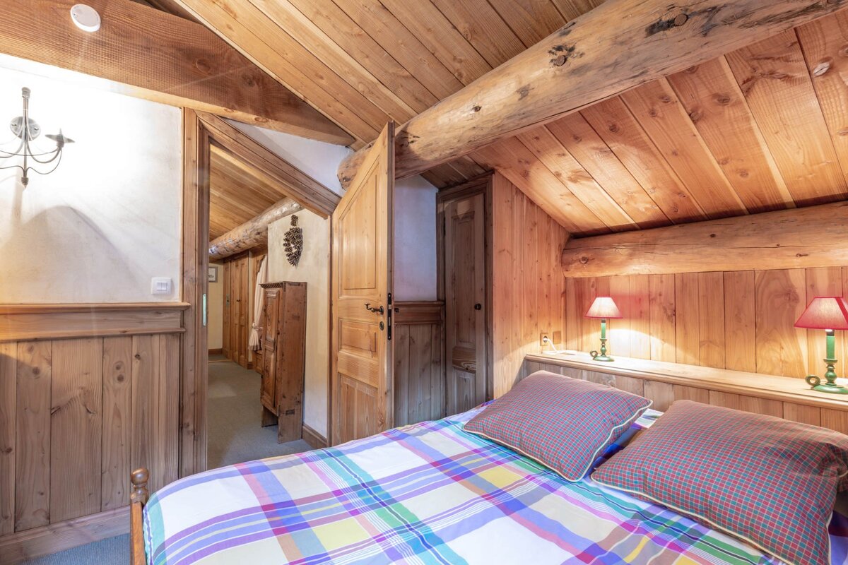A cozy wooden cabin bedroom featuring a bed with a colorful plaid duvet under a vaulted, log-beamed ceiling.