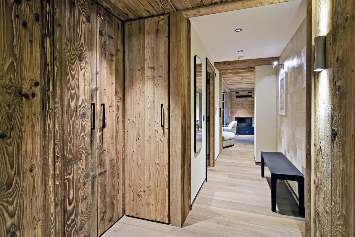 A rustic-modern chalet hallway featuring textured wood paneled wardrobes, a black bench, and a corridor leading to a living area.
