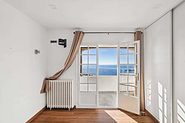 A bright, empty room with white walls, wood floor, and floor-to-ceiling glass doors leading to a balcony with a vast ocean view.