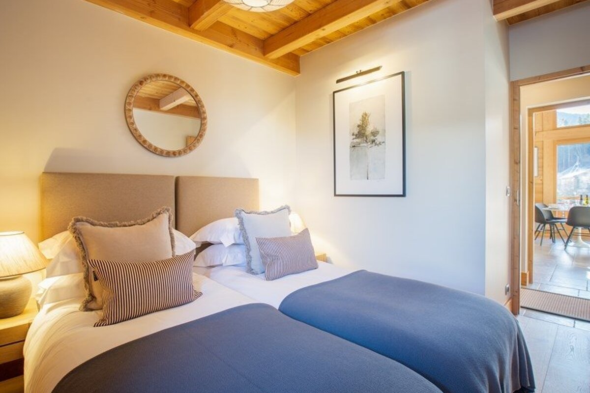 Cozy bedroom featuring two twin beds with blue blankets, a wooden ceiling, a round mirror, and framed artwork.