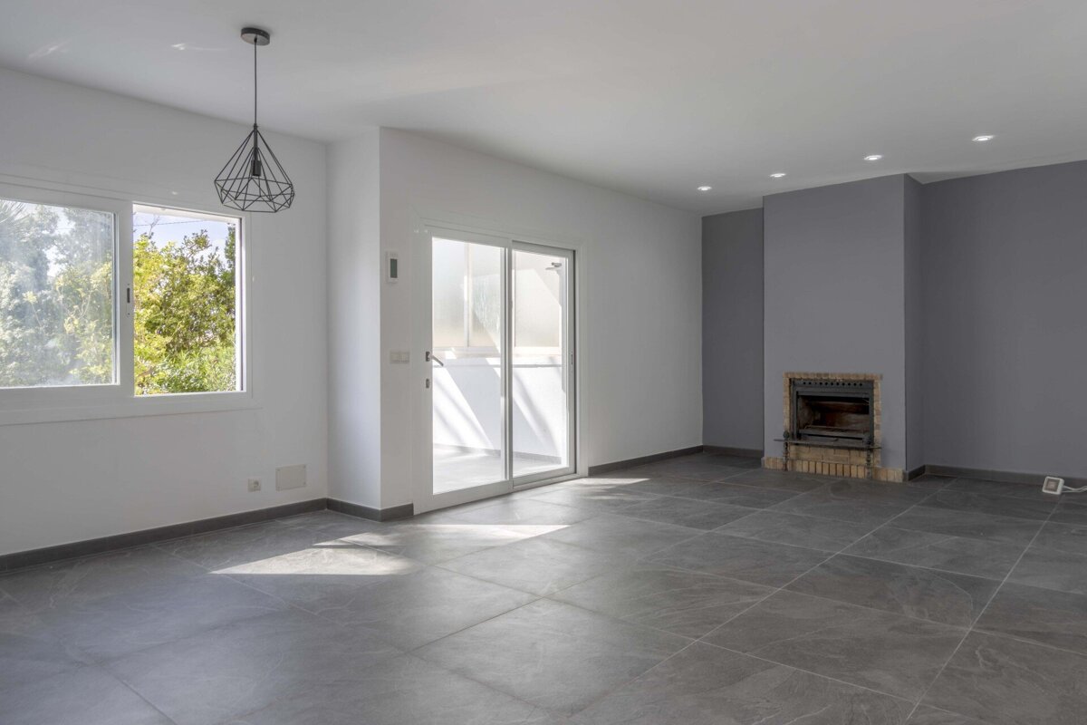 Spacious, empty room with white walls, grey tiled floor, large window, sliding door, a fireplace built into a grey wall, and modern lighting.