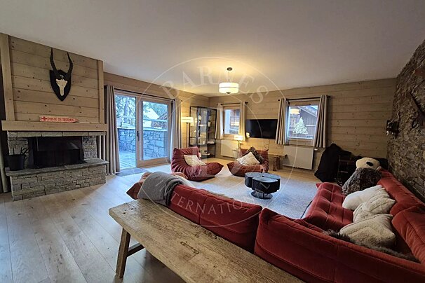An inviting chalet living room with wood-paneled walls, a stone fireplace, red modular sofas, and a large sliding door leading outside.