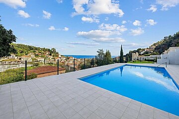 A blue swimming pool on a tiled terrace overlooks a coastal town, the ocean, and green hills under a partly cloudy sky.