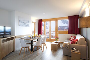A modern living/dining area with a breakfast-set table, a sofa with a Swiss flag pillow, and large windows looking out to a snowy mountain resort.