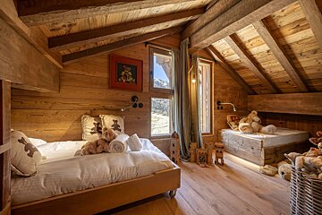 Cozy, rustic wooden attic bedroom with two beds, lots of teddy bears, and windows offering a scenic mountain view.