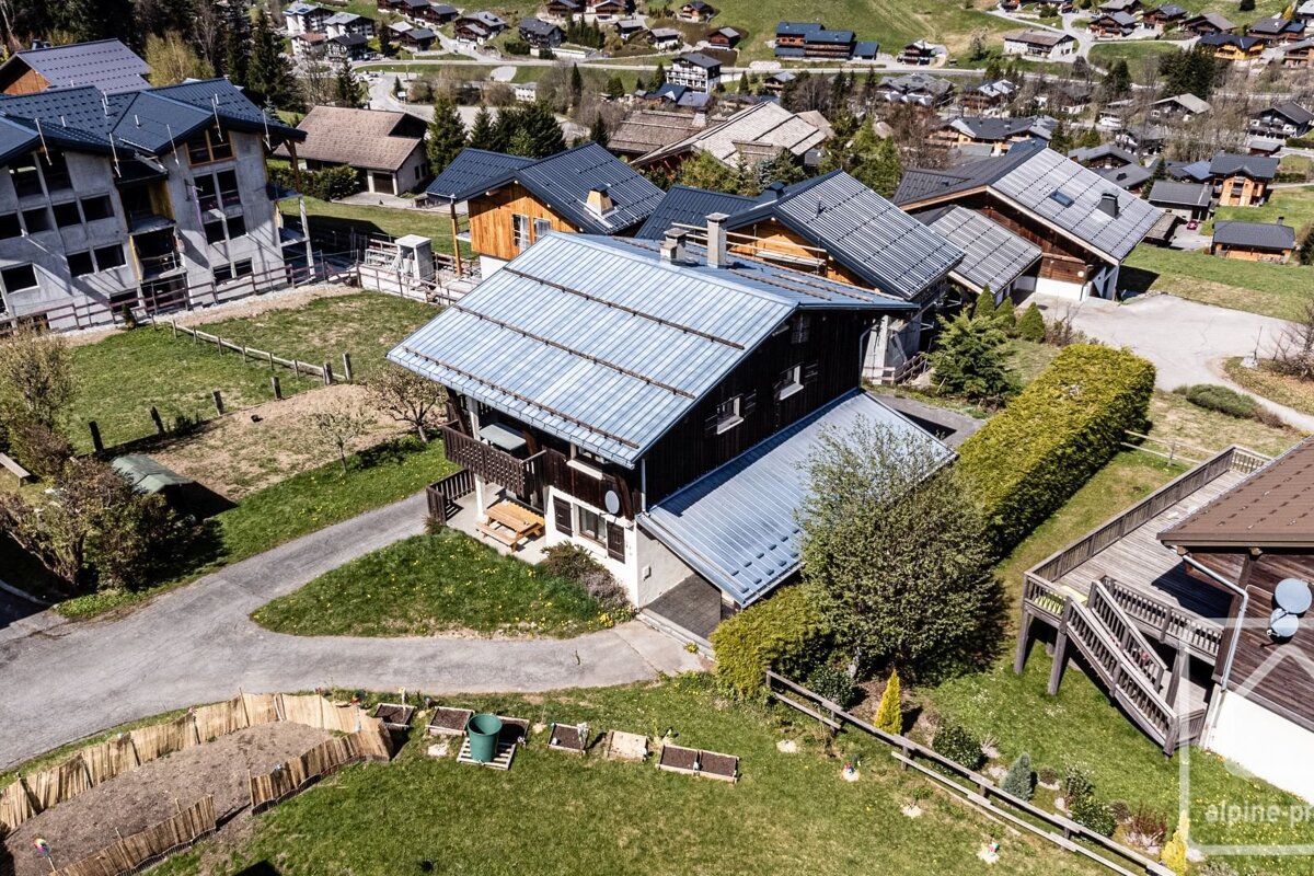 Aerial view of a village with numerous houses, green lawns, and winding roads. A large house with a distinctive metal roof and balcony stands centrally.