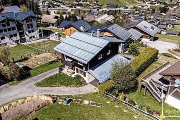Aerial view of a village with numerous houses, green lawns, and winding roads. A large house with a distinctive metal roof and balcony stands centrally.