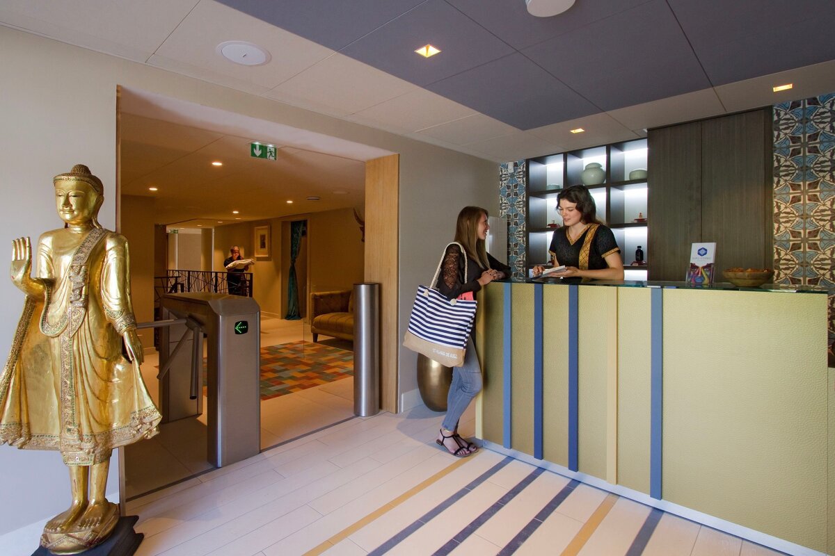 Two women at a reception desk next to a golden Buddha and a turnstile, in a modern, subtly Eastern-themed lobby.