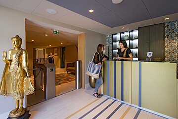 Two women at a reception desk next to a golden Buddha and a turnstile, in a modern, subtly Eastern-themed lobby.