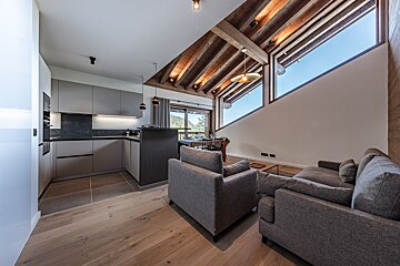 Spacious modern open-plan kitchen, dining, and living area. Features exposed wooden beams, large slanted windows, and grey furnishings.