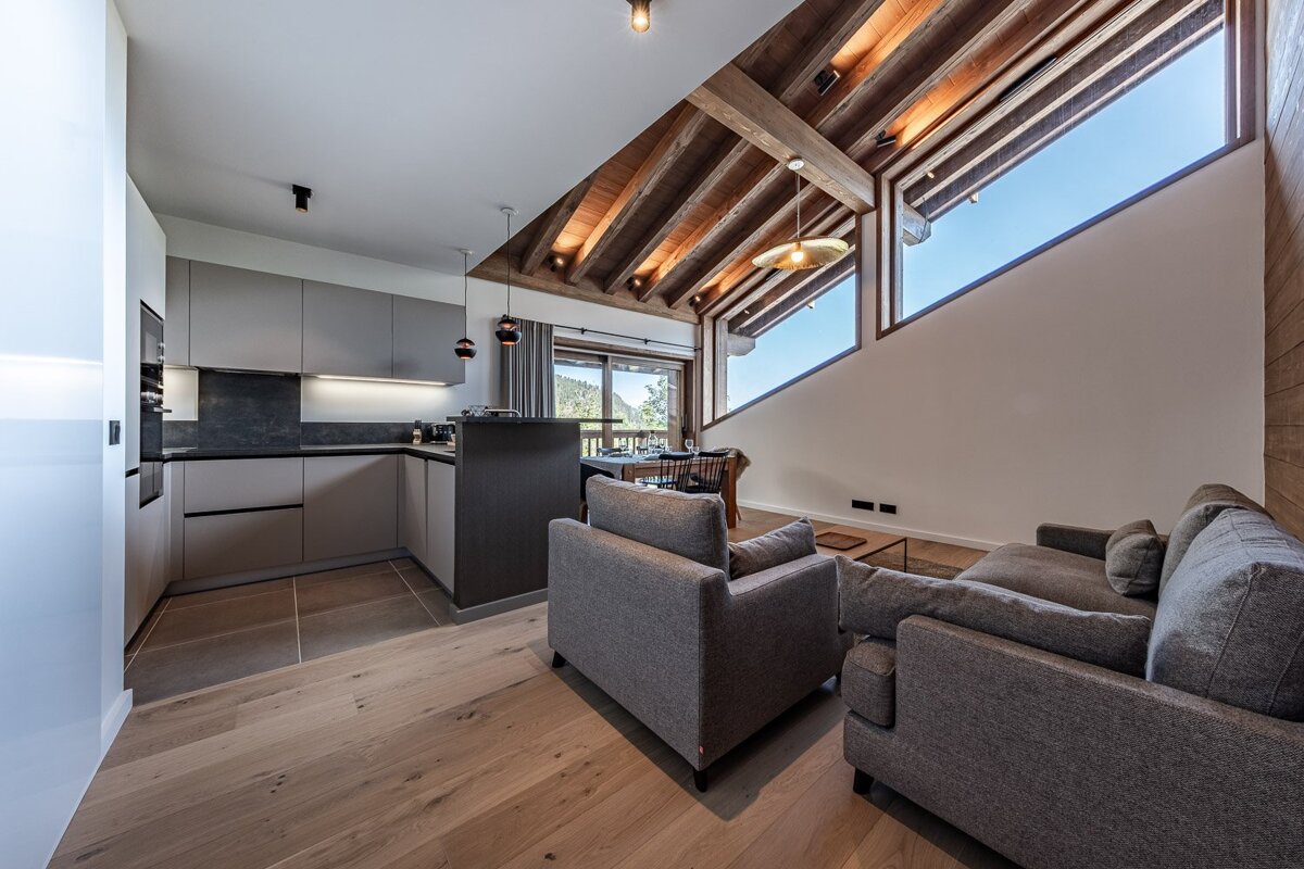 Modern open-plan living area with sleek kitchen, grey sofas, dining, rustic wooden beams, and large windows revealing a scenic mountain view.