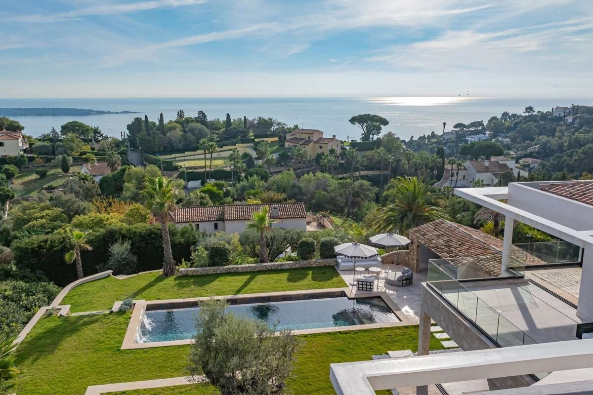 Aerial view of a luxury villa with an infinity pool, manicured lawn, and outdoor lounge, offering stunning ocean views under a clear sky.