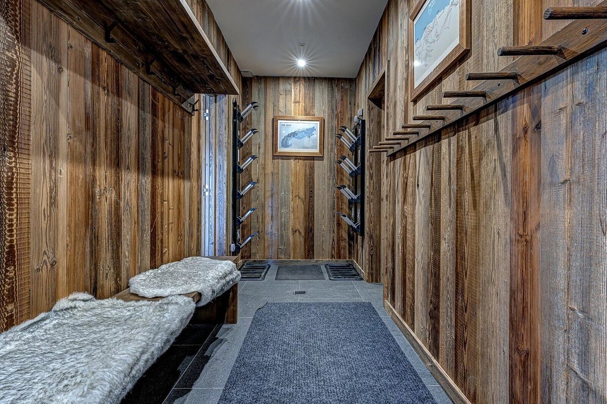 A rustic ski boot room with reclaimed wood walls, ski boot racks, a fur-covered bench, and ski trail maps, creating a cozy chalet atmosphere.