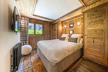 Cozy wood-paneled cabin bedroom featuring a white-dressed bed, wooden wardrobe, rustic rug, and a wall-mounted TV.