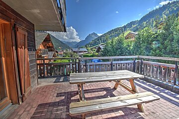 Sunny balcony with a wooden picnic table offers panoramic views of a mountain village, forested valley, and distant peaks under a blue sky.