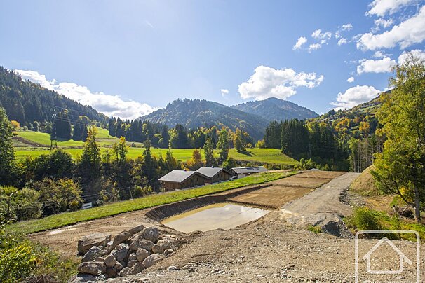 A mountain valley with green hills, forests, and houses. The foreground shows a construction site with dirt, rocks, and a muddy pond under a blue sky.