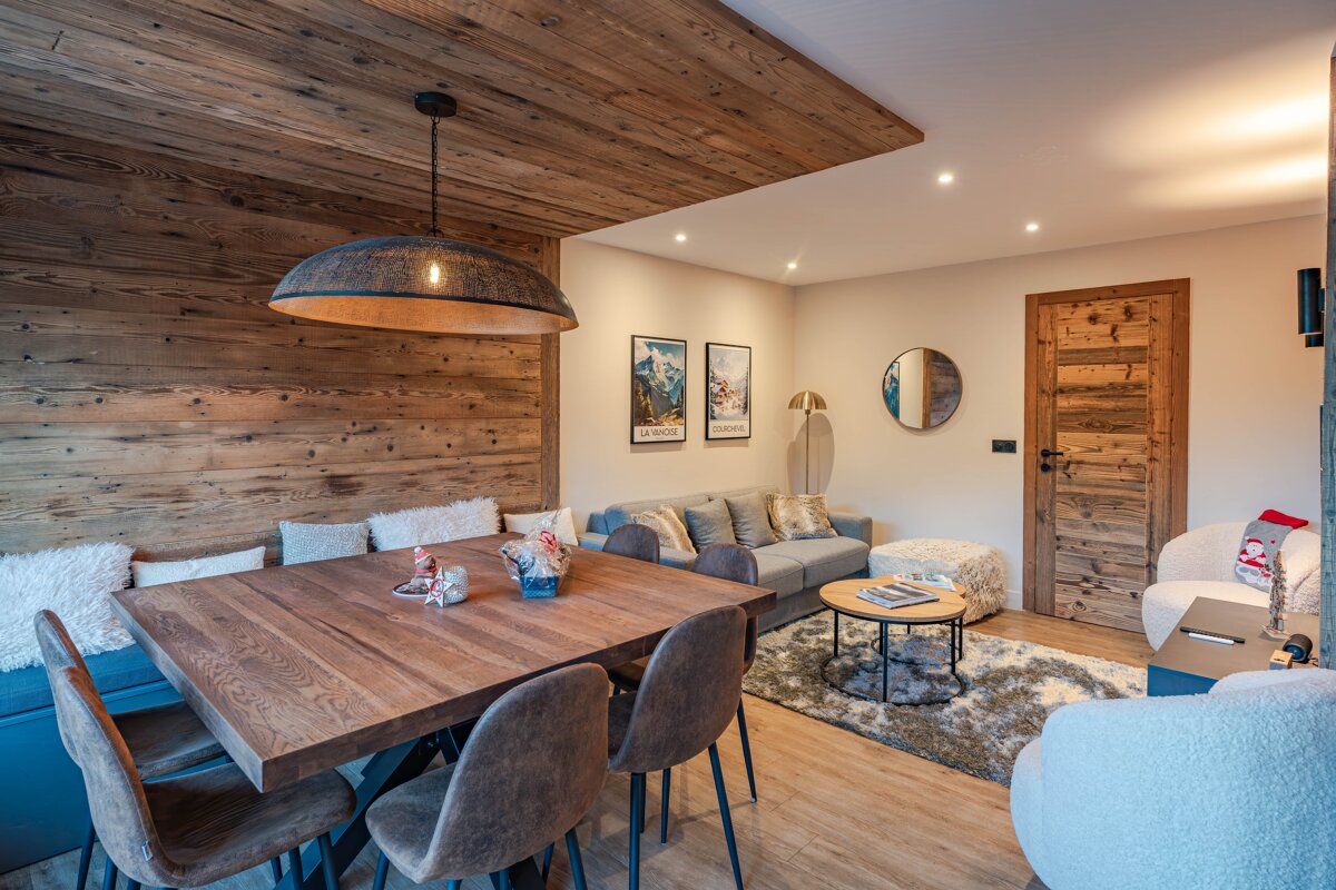 Cozy alpine chalet interior featuring a rustic wood-paneled wall, a dining table, and a living area with a grey sofa and fluffy chairs.