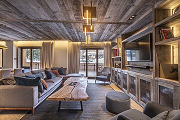 A cozy, rustic living room featuring wooden ceiling/walls, a large sofa, live-edge coffee table, and a snowy outdoor view from the balcony.