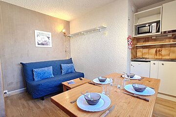 A cozy studio interior featuring a set wooden dining table in the foreground, a blue futon sofa, and a compact kitchenette.