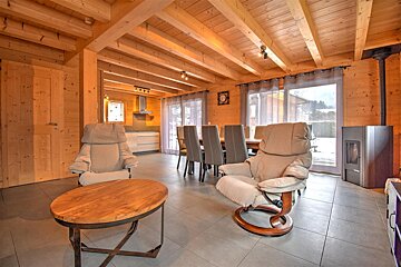 Rustic modern chalet interior with exposed wood, featuring a living area with recliners, dining space, kitchen, wood stove, and large windows.