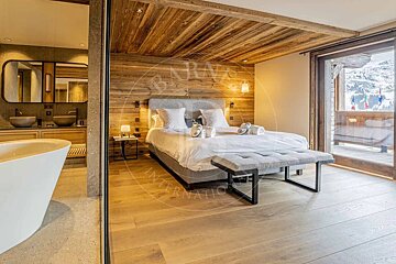 Chic chalet bedroom with wood walls/ceiling, king bed, and open bathroom with tub. A balcony offers snowy mountain views.