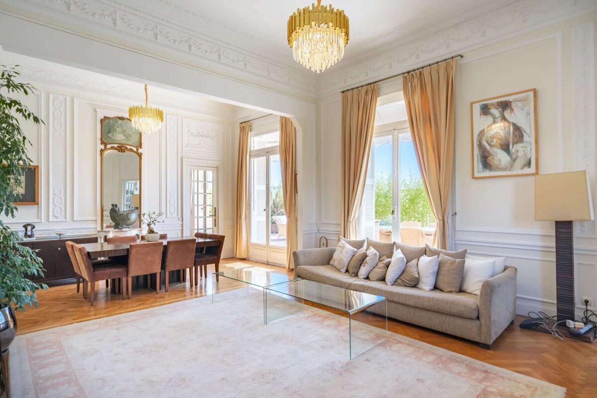 A luxurious, spacious living and dining room with high ceilings, ornate chandeliers, tall windows, and classic decor, filled with natural light.