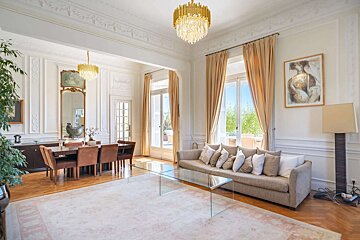 A luxurious, spacious living and dining room with high ceilings, ornate chandeliers, tall windows, and classic decor, filled with natural light.