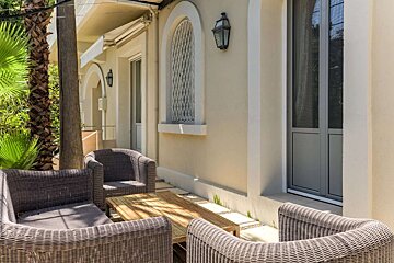 Sunny outdoor patio with wicker furniture and wooden table next to a building with arched windows and palm trees.