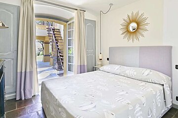 A charming bedroom with open French doors revealing an outdoor staircase and garden. Features a bed with fish-patterned bedding, a sunburst mirror, and lilac accents.