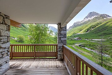 A stone and wood balcony frames a lush green mountain valley with a winding stream, distant peaks, and a small building.