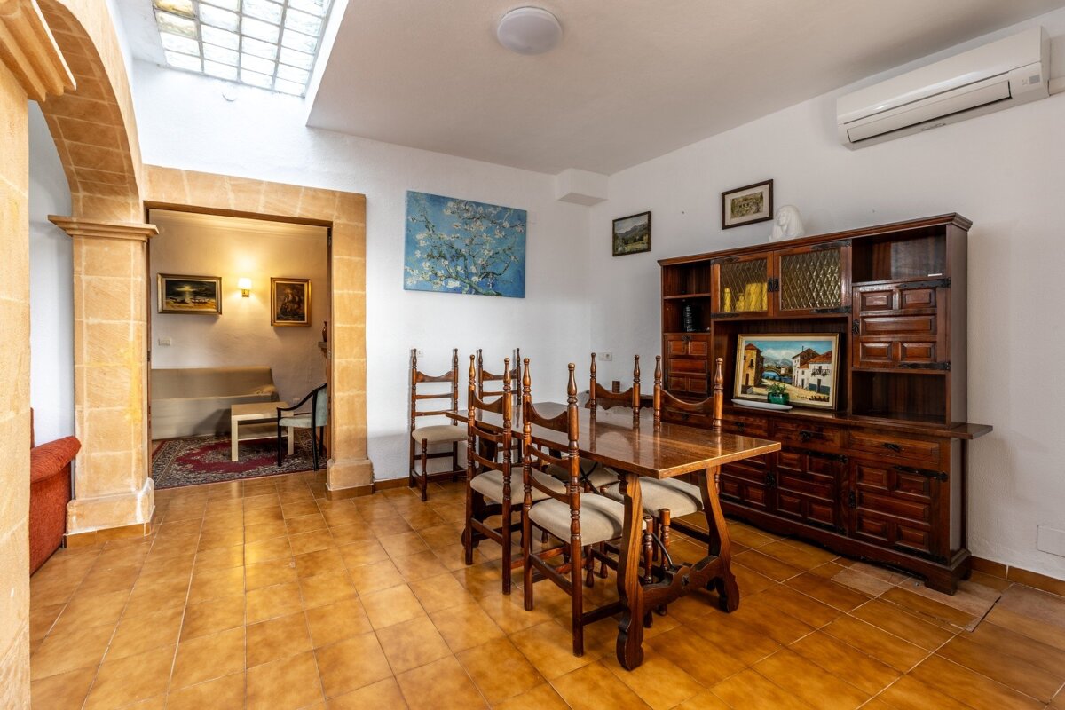 A traditional dining room featuring a long wooden table, chairs, and a large hutch. It has tiled floors, white walls with art, and an archway to a lounge.