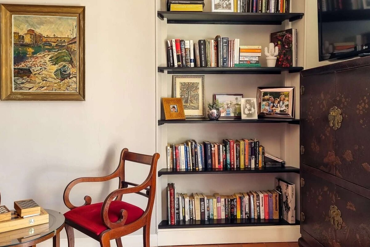A cozy room featuring a tall bookshelf filled with books and framed photos, a vintage red-cushioned chair, a river painting, and an ornate dark cabinet.
