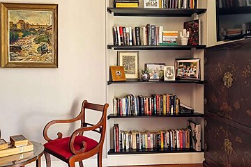A cozy room featuring a tall bookshelf filled with books and framed photos, a vintage red-cushioned chair, a river painting, and an ornate dark cabinet.