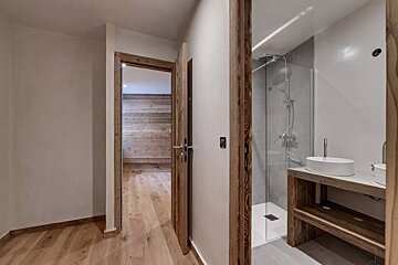 A room with light wood floors and white walls. An open door leads to a wooden room. Another door reveals a modern bathroom with a glass shower and wood vanity.