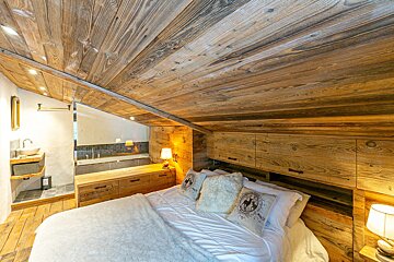 A rustic, cozy bedroom features a white bed, extensive reclaimed wood paneling on the ceiling and walls, and an open-concept bathroom with a sink and tub.