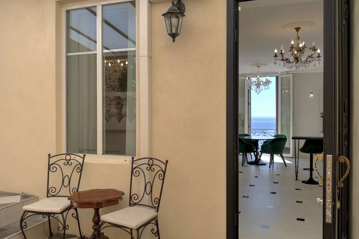 An inviting outdoor area with seating, next to a window, and an open door revealing an elegant room with chandeliers and a panoramic sea view.
