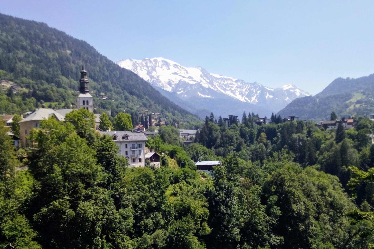 Alpine village with a church spire nestled among lush green forests, against a backdrop of towering snow-capped mountains.