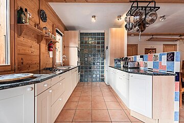 Rustic wood-paneled kitchen with white cabinets, dark counters, a glass block wall, and hanging pots. Features a colorful tiled backsplash.