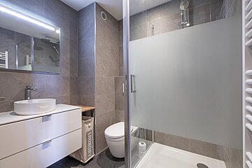 Modern grey-tiled bathroom with a white vanity, basin, toilet, and a walk-in shower with a frosted glass screen. A laundry hamper is visible.
