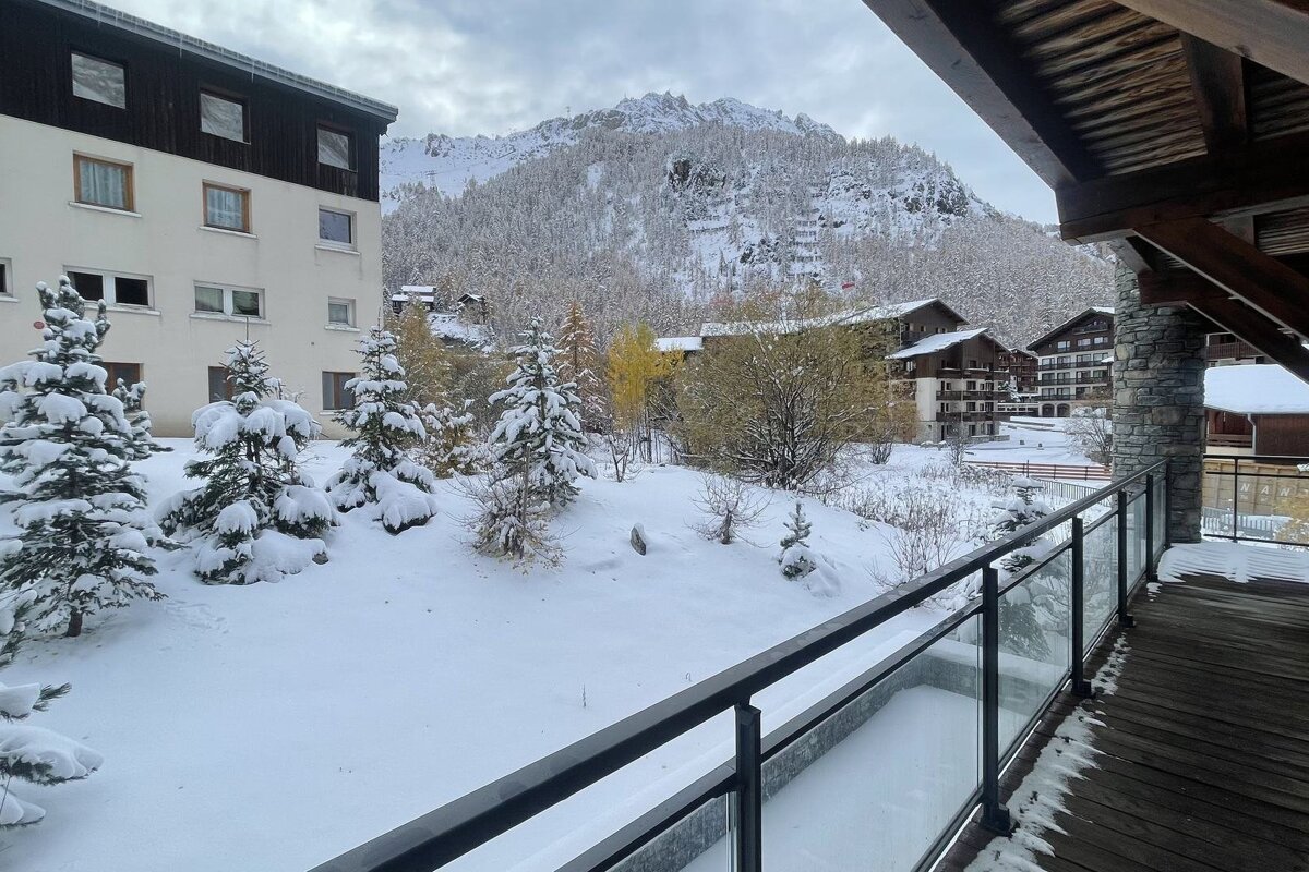 A balcony view of a snowy mountain village with snow-laden trees, distant peaks, and resort buildings under a gray sky.
