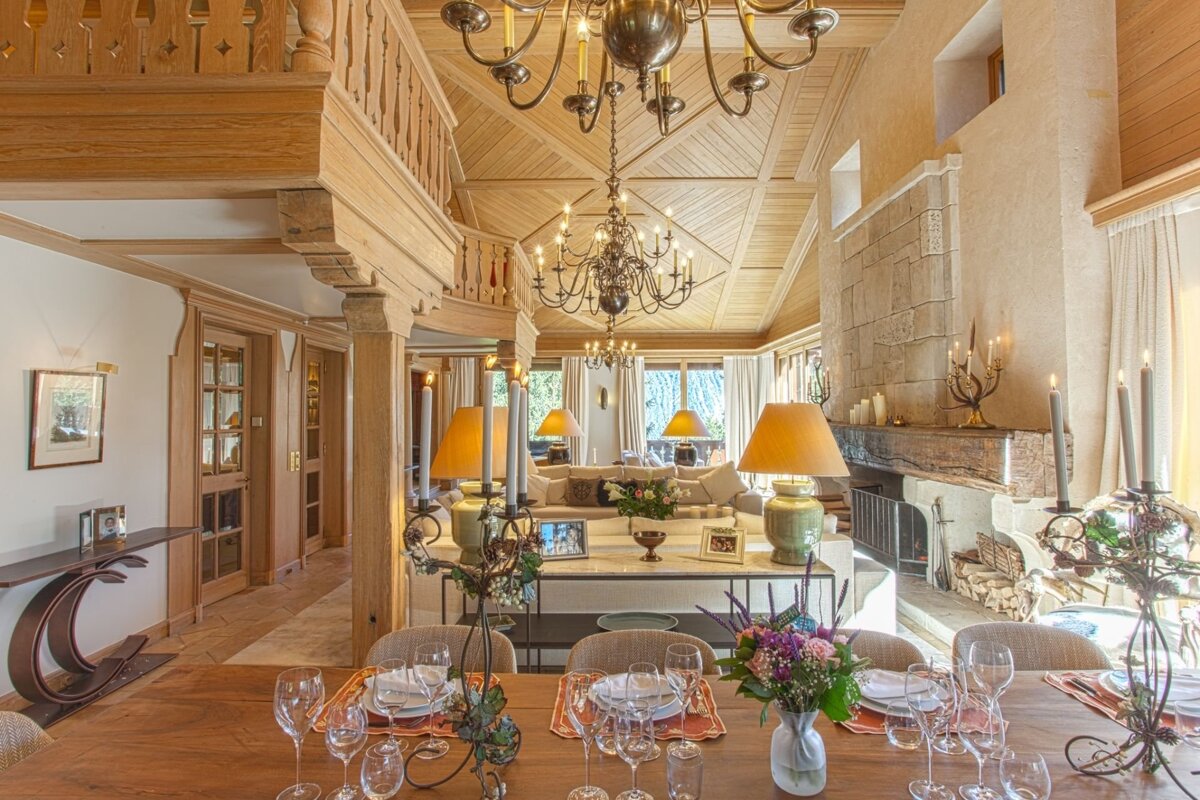 A grand, rustic chalet interior with a set dining table in the foreground, a cozy living area, stone fireplace, and ornate wooden ceilings with chandeliers.