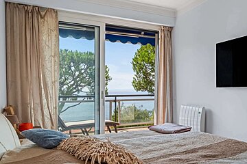 A cozy bedroom with a bed in the foreground, facing an open balcony that offers a stunning view of the sea and lush pine trees.