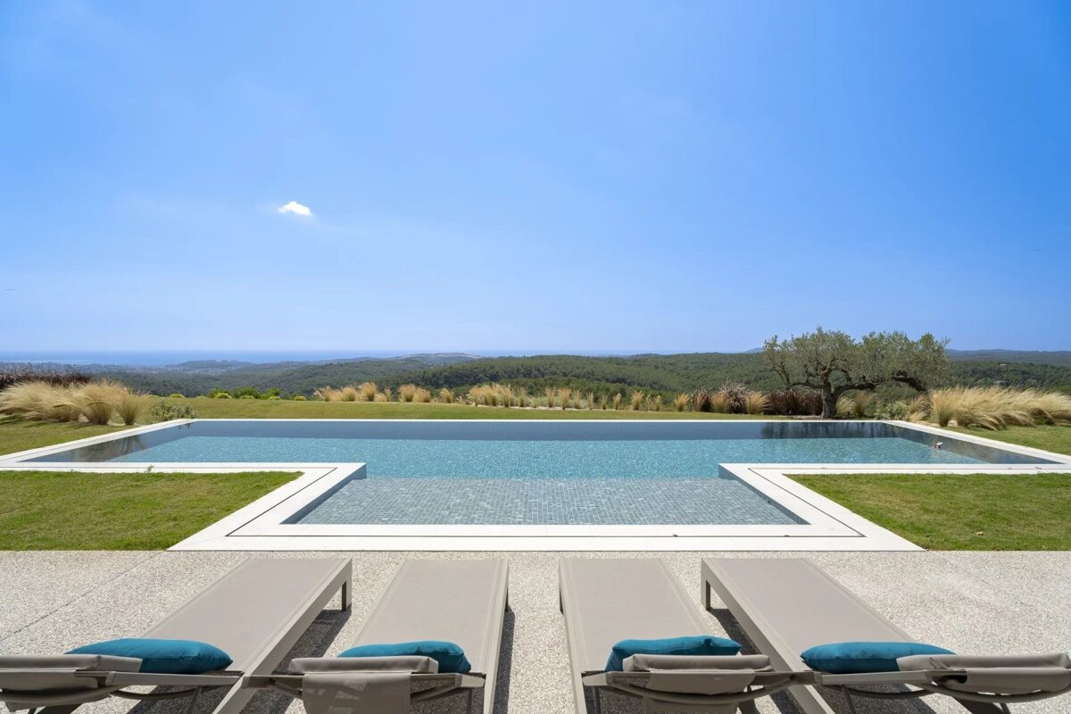 An infinity pool overlooking lush green hills and the ocean under a clear blue sky, with four lounge chairs in the foreground.