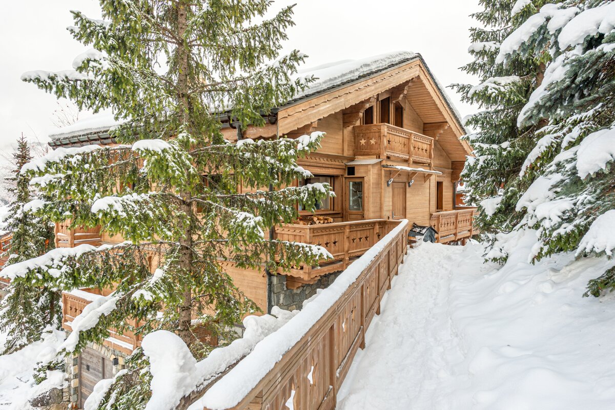 A wooden chalet blanketed in snow, nestled among snow-covered pine trees with a snowy path leading up to it.
