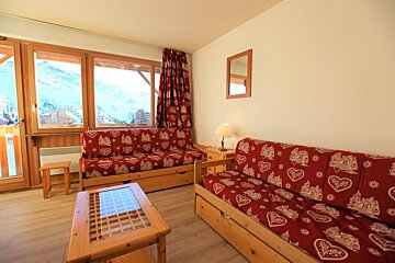 Cozy ski resort apartment living room with two red patterned sofa beds, wooden furniture, and large windows revealing a snowy mountain view.